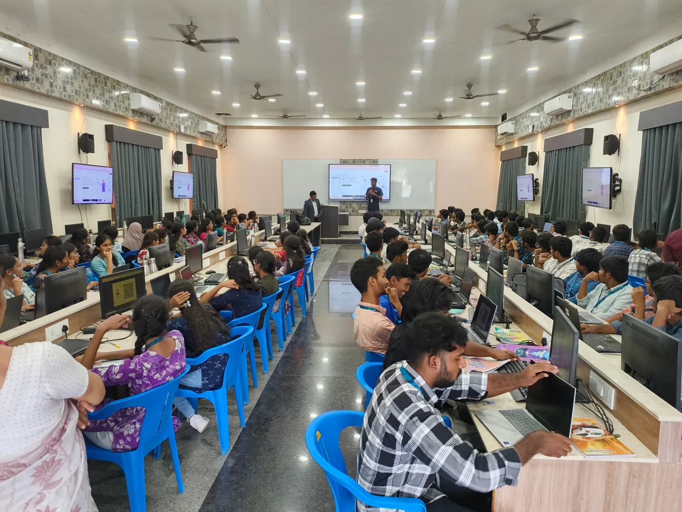 Students at rows of desktop computers facing a shared screen during a classroom session.